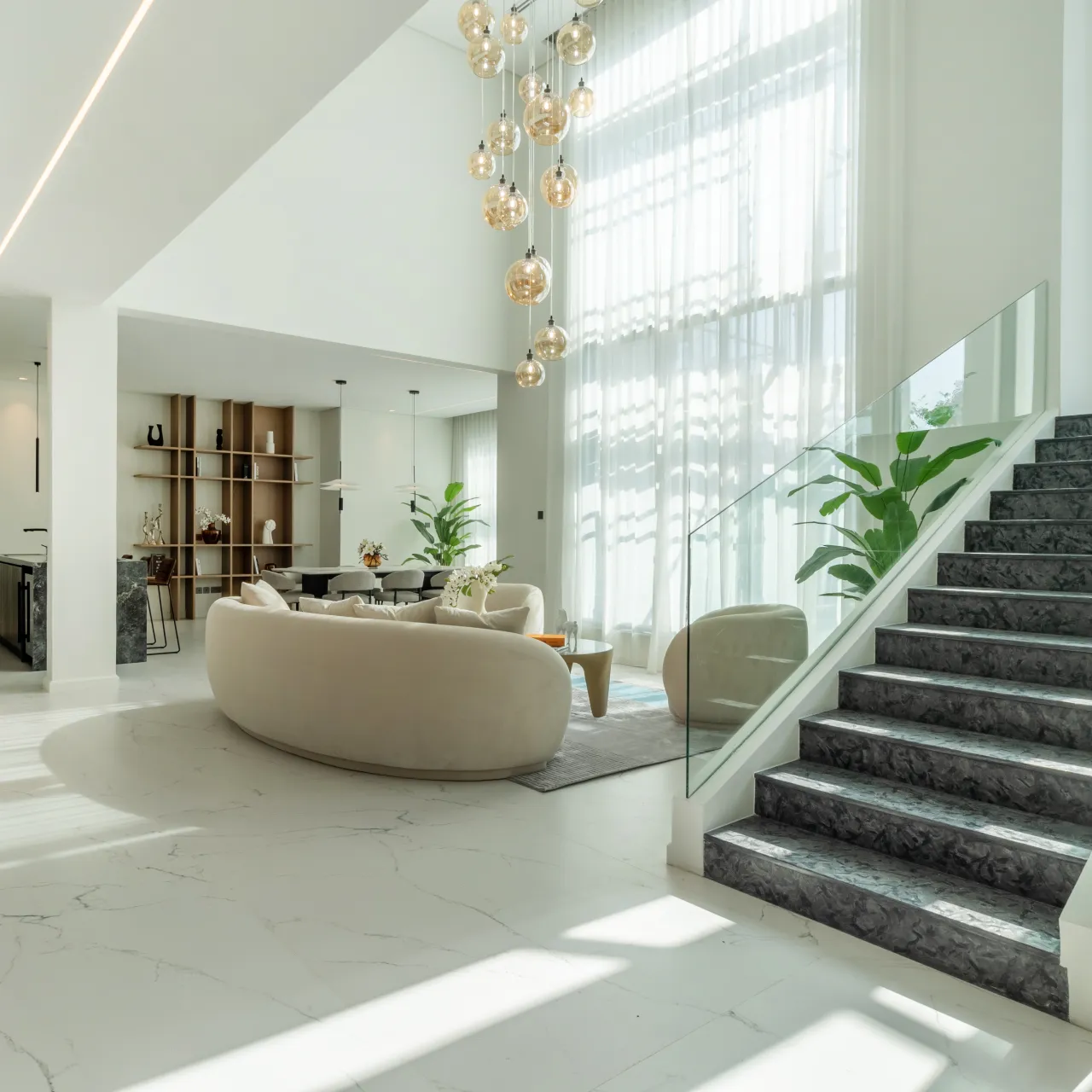 Modern house renovation featuring a bright double-height living room with a curved cream sofa, marble flooring, glass staircase, custom shelving, tall sheer curtains, indoor plants, and warm globe pendant lights.